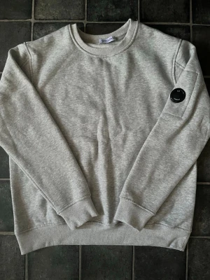 C.P. Company sweatshirt - Snygg grå sweatshirt från C.P. Company med klassisk rund halsringning och ribbade muddar. Perfekt för en avslappnad och stilren look.          OBS: Tröjan är nopprig på insidan och släpper av väldigt mycket smular, den är köpt på marknad så vet ej om autenticiteten.