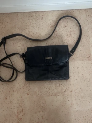 Svart axelväska i skinn från 7EEL - Snygg svart axelväska i äkta skinn från Face. Väskan har justerbar axelrem, lock med magnetknäppning och flera fack med dragkedja. Perfekt storlek för det viktigaste och stilren metallbricka framtill.