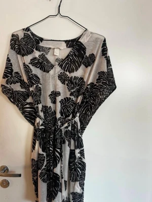 Svartvit blommig tunika - Snygg svartvit tunika med stora blommönster och omlottknytning i midjan. Klänningen har v-ringning och vida, korta ärmar. Perfekt för dig som gillar en avslappnad men trendig stil. Rök och djurfritt hem. Pruta gärna 🌸den är stor i stoleken!
