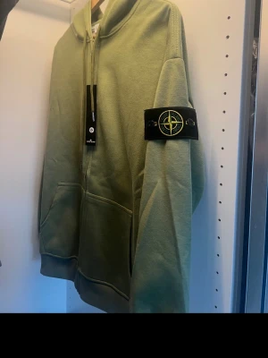 Grön hoodie från Stone Island - Snygg ljusgrön hoodie från Stone Island med dragkedja framtill och klassisk logopatch på vänster ärm. Hoodien har två fickor fram och ribbade muddar. Tillverkad i mjukt bomullsmaterial och har en avslappnad passform. Perfekt för en streetwear-look.