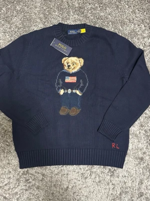 Mörkblå stickad tröja Polo Bear - Mörkblå stickad tröja från Polo Ralph Lauren med ikoniska Polo Bear-motivet på bröstet. Tröjan har rund hals, ribbade muddar och RL-broderi vid nederkanten. Materialet är mjukt och skönt, perfekt för kyligare dagar.