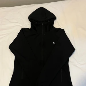 Svart tränings tröja  - Svart tränings tröja från Haglöfs med hel dragkedja, två fickor med dragkedja och justerbar huva. Tröjan har en stilren look och är perfekt för att träna med. Materialet är lätt och smidigt, vilket gör den enkel att bära med sig. Klassisk passform och långärmad.