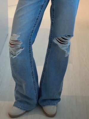 Ljusblå slitna bootcut jeans - Säljer ett par ljusblå bootcut jeans med stora slitningar över knäna och klassiska bakfickor. Jeansen har en avslappnad passform och är tillverkade i denim. Perfekta för en trendig och avslappnad stil.