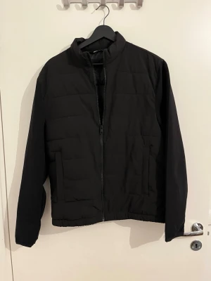 Svart pufferjacka från Zara - Säljer en svart pufferjacka från Zara med hög krage och dragkedja framtill. Jackan har två sidofickor och quiltad design som ger en clean och modern look. Perfekt för kalla vinterdagar tack vare det vadderade materialet.