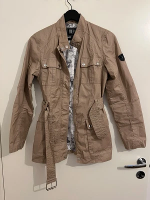 Beige jacka från Jofama - Stilren beige utilityjacka från Jofama med bälte i midjan, fyra framfickor med tryckknappar och ståkrage. Jackan har ett coolt mönstrat innerfoder och patch på ärmen. Perfekt för dig som gillar en avslappnad men trendig look.