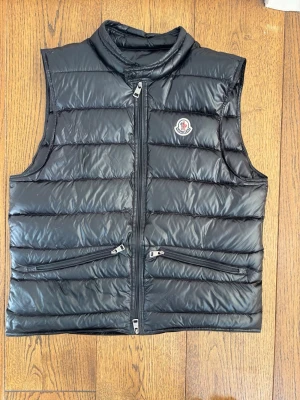 Svart dunväst från Moncler storlek 2 - Hör av er vid frågor! Snygg svart dunväst från Moncler med quiltad design och hög krage. Västen har två dragkedjeförsedda fickor framtill och klassisk Moncler-logga på bröstet. Perfekt för lager-på-lager och ger en clean look till din outfit.