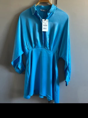Blå skjortklänning från Zara - Snygg blå skjortklänning från Zara i ett glansigt material. Klänningen har krage, knappar framtill och trekvartsärm med knapp vid ärmslut. Perfekt för dig som vill ha en stilren och trendig look.