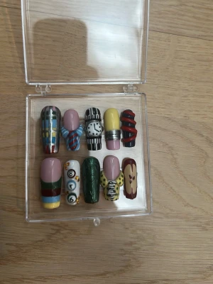 Unika press-on naglar med 3D-design - Säljer ett set press-on naglar med handmålade 3D-motiv i olika färger och former. Naglarna har detaljer som klocka, slips och rutiga mönster i nyanser av grön, gul, röd, svart och blå. Perfekt för dig som vill sticka ut med kreativa och färgstarka naglar.  Tumme: 13mm pek: 10mm lång: 12mm ring: 10mm Lill: 8mm