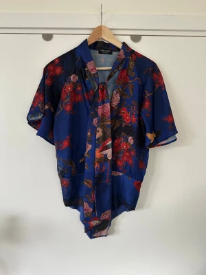 Blommig blå blus / body från Zara - Superfin blå mönstrad blus / body / topp från ZARA i storlek S med knytning i halsen och vida korta ärmar. Färgglatt mönster av detaljerade blommor, fjärilar och fåglar i ett lent halvglansigt satin-material. Praktiska tryckknappar i grenen, vilket ger en slät passform. Fin!