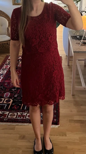 Spetsklänning  - Vinröd klänning i spets med rund halsringning och korta ärmar. Klänningen har en figurnära passform och slutar ovanför knät. Spetsdetaljerna ger en romantisk känsla och klänningen har en mjuk underklänning för extra komfort.