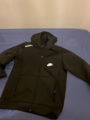 Svart Nike Tech Fleece hoodie - Säljer en svart Nike Tech Fleece hoodie med dragkedja och huva. Hoodien har vita Nike-loggor på bröstet och axeln, samt två fickor med dragkedja framtill. Tillverkad i mjukt och stretchigt material som känns riktigt skönt mot huden. Fickornas dragkedja funkar inte dock 