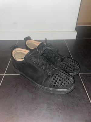 Svarta sneakers med nitar - Snygga helsvarta sneakers från Christian Louboutin med mattsvarta nitar på tån och svart mocka. Klassisk låg modell med svarta skosnören och gummisula. Perfekta för dig som vill sticka ut med en edgy look.