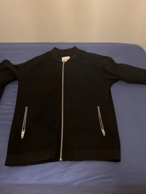 Svart bomberjacka från Jack & Jones - Snygg svart bomberjacka från Jack & Jones med silverfärgade dragkedjor framtill och på fickorna. Klassisk ribbad krage och muddar. Tillverkad i ett mjukt material som ger en clean och stilren look.