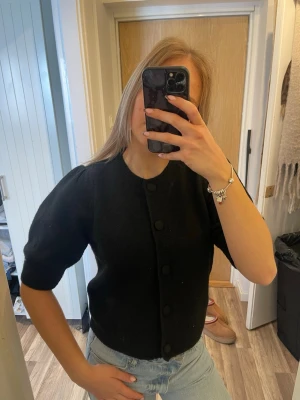 Svart puffärmad kofta - Snygg svart kofta med puffärmar och stora knappar framtill. Kortare modell som passar perfekt till höga jeans. Koftan har rund halsringning och är gjord i ett mjukt stickat material.