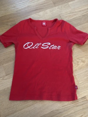 Y2K Röd Converse All Star t-shirt - Snygg röd t-shirt från Converse All Star med vit broderad text på bröstet. T-shirten har korta ärmar, unik v-ringad halsringning och är gjord i mjuk bomull. Perfekt för en avslappnad och sportig stil.
