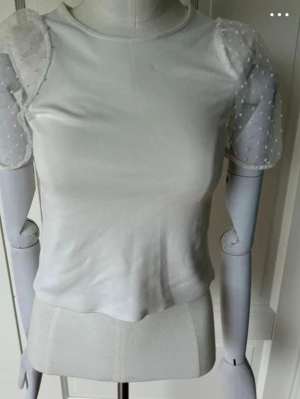 Vit topp med puffärmar i mesh - Söt vit topp med transparenta puffärmar i mesh och diskreta prickar. Kroppsdelen är i mjuk bomullsmix och toppen har en figurnära passform. Säljer pga att den inte passar och storleken kan upplevas som lite större än vad det står💓