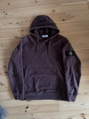 Brun hoodie från Stone Island - Säljer en brun hoodie från Stone Island med klassisk patch på vänster ärm. Hoodien har huva med dragsko och en stor magficka framtill. Tillverkad i mjuk bomull för skön känsla och avslappnad stil. Perfekt för dig som gillar streetwear och vill ha något stilrent. Hoodien har inga defekter, helt äkta till 100%