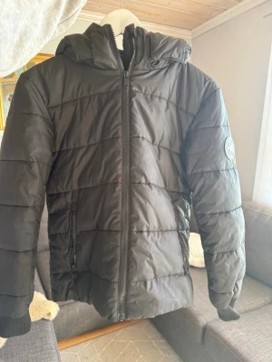 Svart pufferjacka från Everest - Snygg svart pufferjacka från Everest med huva och dragkedja framtill. Jackan har två fickor med dragkedja och ribbade muddar vid ärmsluten. Märke med Everest-logga på ena ärmen. Perfekt för kalla vinterdagar.