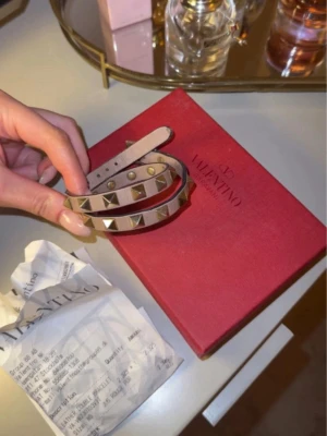 Valentino Rockstud armband beige - Snyggt Valentino Rockstud armband i beige läder med dubbla rader och ikoniska guldfärgade nitar. Justerbar längd med flera hål för perfekt passform. Kommer med original ask och kvitto. Perfekt accessoar för att lyfta din outfit. Pris kan diskuteras 