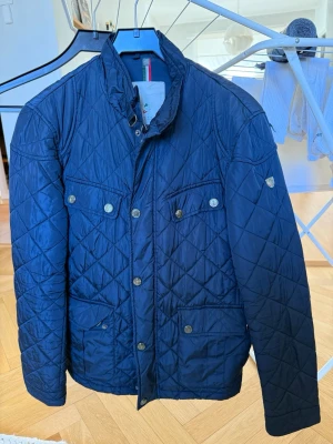 Mörkblå quiltad jacka med knappar - Snygg mörkblå quiltad jacka med hög krage och flera fickor framtill. Jackan har tryckknappar och dragkedja, samt ett klassiskt rutmönster. Perfekt för vårdagar och ger en stilren look.