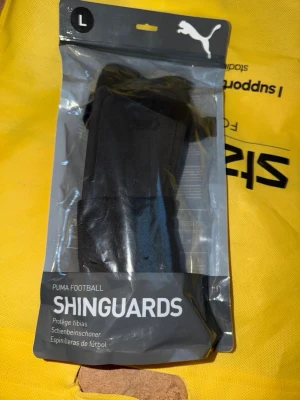 Puma benskydd svart storlek L - Säljer ett par svarta benskydd från Puma, modell Shinguards, i storlek L. Perfekta för fotbollsträning eller match. De har en klassisk design med elastiska band för att sitta på plats och skydda smalbenen. Kommer i originalförpackning.