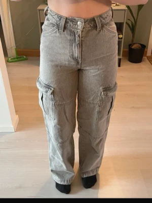 Grå cargobyxor med raka ben - Säljer ett par grå cargobyxor i denim med raka ben och stora sidofickor. Byxorna har hög midja. Perfekta för en avslappnad och trendig look. Materialet är kraftigt och ger en cool vintagekänsla.