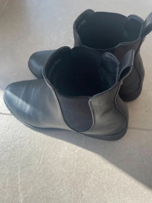 Svarta Chelsea boots i skinn - Stilrena svarta Chelsea boots i slätt skinn med klassiska resårpaneler på sidorna och rundad tå. Sitter på låg sula och har dragflik baktill för enkel påtagning. Perfekta för en trendig och tidlös look.