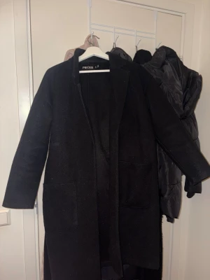 Svart  kappa från Nelly - Stilren svart lång kappa från Nelly med raka ärmar och två stora fickor framtill. Kappan har ett bälte i midjan för att kunna knyta och skapa en snygg siluett. Perfekt för kyligare dagar och enkel att matcha med olika outfits. Har använts några gånger men ser ut som ny.