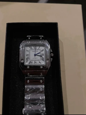  herrklocka i silver - Säljer en stilren herrklocka i silverfärgat stål med fyrkantig boett, romerska siffror och blå visare. Klockan har en länkad armlänk med synliga skruvar och automatisk urverk. Perfekt för dig som gillar klassisk och lyxig design.