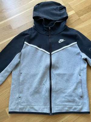 Nike hoodie i svart och grått. Strl xl(barn storlek) - Nike hoodie med dragkedja i svart och ljusgrått. Snygg vit detalj över bröstet och klassisk Nike-logga på vänster sida. Huva och två fickor framtill. Tillverkad i mjukt sweatshirtmaterial, perfekt för chill eller träning.