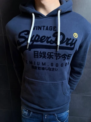 Blå Superdry hoodie med tryck - Mörkblå hoodie från Superdry med stor logga och japanskt tryck på bröstet. Klassisk känguruficka framtill, vita dragsnören i huvan och ribbade muddar. Perfekt för en avslappnad stil. Mjuk bomullsblandning för extra komfort. Hör gärna av dig vid minsta fundering!