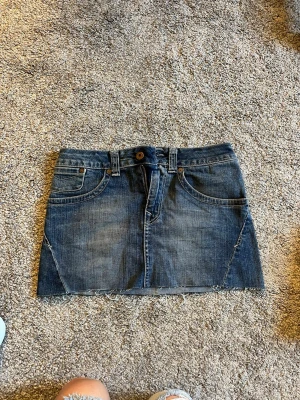 Blå jeanskjol från crocker - Säljer en klassisk blå jeanskjol från crocker med rå kant nertill och femficksmodell. Kjolen har bälteshällor, knapp- och dragkedjestängning samt snygga bakfickor med broderade detaljer. Perfekt för en avslappnad och trendig look.