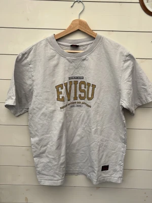 Y2K Evisu grå t-shirt med tryck - Snygg vit t-shirt från Evisu med stort guldfärgat tryck på bröstet och klassisk rund hals. Tillverkad i mjuk bomull och har en relaxed fit med korta ärmar. Perfekt för dig som gillar streetwear och vill ha något stilrent men ändå unikt.