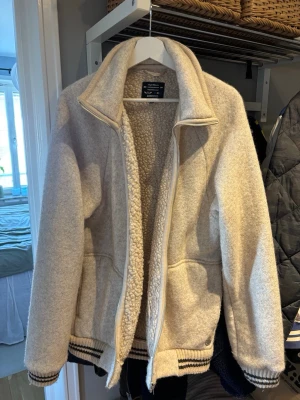Peak Performance Beige jacka med dragkedja - Mysig beige teddyjacka från Peak Performance med mjukt, fluffigt foder och bred krage. Jackan har dragkedja framtill, två fickor och ribbade muddar med svarta och vita ränder. Perfekt för kyliga dagar och ger en avslappnad, trendig look.