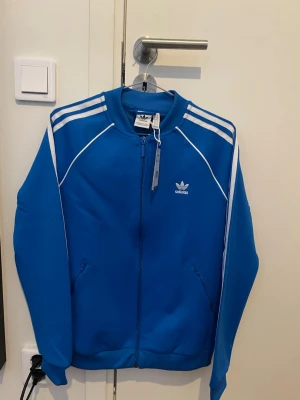 Blå Adidas ziptröja med vita ränder - Snygg blå ziptröja från Adidas med klassiska vita ränder längs ärmarna och Adidas-logga på bröstet. Tröjan har dragkedja framtill, två fickor och ribbade muddar. Perfekt för en sportig och avslappnad stil.