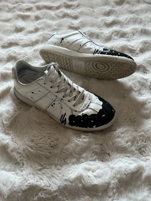 Maison Margiela sneakers svart/vit - Maison Margiela sneakers i vitt läder med svart färgstänk och målad tå. Klassisk låg modell med vita snören och gummisula. Unik design med konstnärlig touch, perfekt för dig som vill sticka ut. Man får med boxen, dem är använda men fortfarande i bra skick. Priset är diskuterbart.