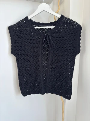 Svart virkad väst med knytband - Snygg svart stickad väst med virkat hålmönster och knytband framtill. Västen har korta ärmar och ribbade kanter runt hals och ärmslut. Perfekt att slänga över en t-shirt eller skjorta för en trendig lager-på-lager-look.