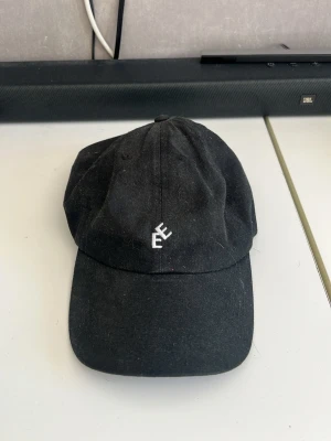 Svart keps från Eytys med broderad logga - Säljer en svart keps från Eytys med böjd skärm och justerbar rem bak. Kepsen har en stilren vit broderad Eytys-logga framtill och är tillverkad i bomull. Perfekt accessoar för en clean och avslappnad look.