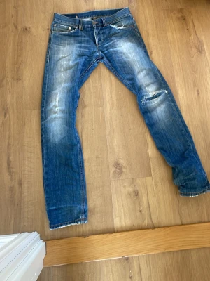 Blå slitna jeans med hål - Säljer ett par blå jeans med slitningar och hål på knät. Jeansen har klassisk femficksmodell, normal midja och raka ben. Snyggt tvättade detaljer och slitna kanter för en avslappnad look. Perfekta för dig som gillar en trendig och casual stil.