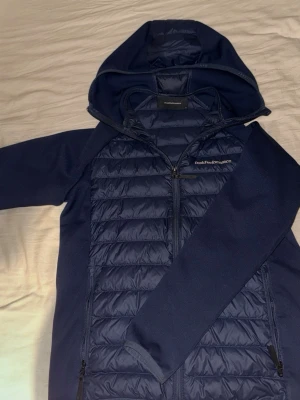 Mörkblå cardigan från Peak Performance - Snygg mörkblå cardigan från Peak Performance med vadderade paneler fram och bak, huva och hel dragkedja. Jackan har två sidofickor med dragkedja och är tillverkad i en mix av syntetmaterial och quiltat tyg för extra värme och stil. Perfekt för vädret just nu