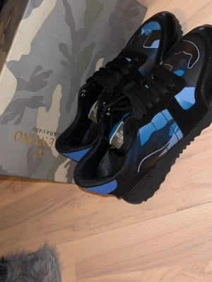 Valentino Garavani blå camo sneakers - Snygga sneakers från Valentino Garavani med blå och svart camo-mönster. Skorna har svarta snören, svarta detaljer i mocka och läder samt coola nitar på sulan. Perfekt för dig som vill sticka ut med en unik design.