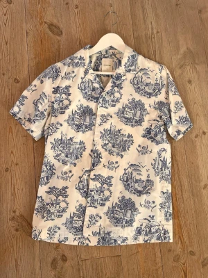 Wood Wood Skjorta - Säljer en vit kortärmad skjorta från Arket med blått, detaljerat mönster över hela plagget. Skjortan har klassisk krage och knappar framtill. Perfekt för dig som gillar unik stil och vill sticka ut lite extra.