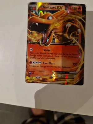 Charizard EX Pokémonkort - Säljer ett Charizard EX samlarkort från Pokémon TCG. Kortet har en glansig yta med färger i orange, svart och guld. Motivet visar Charizard omgiven av eld och kortet har attacker som Stoke och Fire Blast. Perfekt för dig som gillar att samla eller spela Pokémonkort.