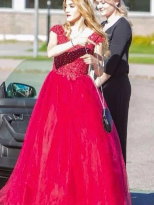 Röd/vinröd balklänning - Säljer en röd långklänning med v-ringning, dekorerad med spets och pärlor upptill. Kjolen är vid och i flera lager för en extra volym. Klänningen har korta ärmar och passar perfekt till bal eller festliga tillfällen. Passar perfekt till 34-36 eventuellt 38 också. Använd endast en gång.