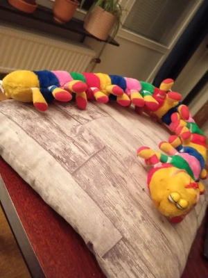 Färgglad mjukisdjur tusenfoting - Säljer en lång och mjuk tusenfoting i plysch med färgglada ränder i gult, blått, grönt, rosa och rött. Den har många ben och ett gulligt ansikte med broderade ögonfransar och rosett. Perfekt som dekoration eller kramdjur.