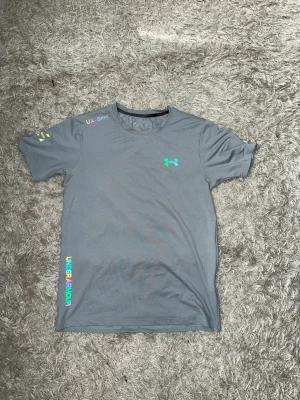 Grå Under Armour t-shirt (reflex) - Snygg grå t-shirt från Under Armour med färgglada loggor och textdetaljer på bröstet (reflex), axeln och sidan. Tillverkad i ett mjukt och lätt material som passar perfekt till träning. Klassisk rund halsringning och korta ärmar. Hela settet finns i min profil som en egen annons.