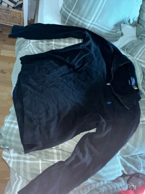 Svart halvzip från Polo Ralph Lauren - Klassisk svart tröja från Polo Ralph Lauren med halv dragkedja och diskret broderad logga på bröstet. Tillverkad i bomull med ribbade muddar och krage. Perfekt för en stilren och avslappnad look. Köpt från tj max i Usa, passar S/M