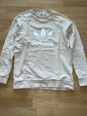 Beige sweatshirt från Adidas - Säljer en beige sweatshirt från Adidas med det klassiska Trefoil-loggan tryckt i vitt framtill. Tröjan har rund halsringning, långa ärmar och ribbade muddar. 