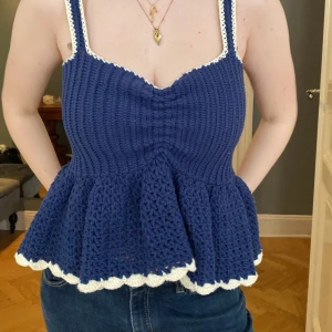 Blå virkad linnetopp med volang - Supersöt blå handvirkad linnetopp med volangkant och vita detaljer längs kanterna. Toppen har smala axelband och en figurnära passform upptill som blir vidare nedtill. Perfekt för sommarens alla tillfällen och ger en trendig, bohemisk vibe.