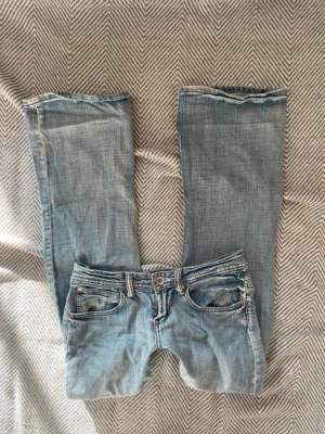 Ljusblå bootcut jeans - Säljer ett par ljusblå bootcut jeans med klassisk femficksdesign och slitna detaljer. Jeansen har lågmidja och ljus tvätt, perfekta för en avslappnad och trendig look. 
